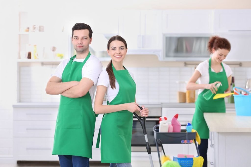 Three individuals wearing green aprons stand together in a kitchen, surrounded by cooking utensils and ingredients.