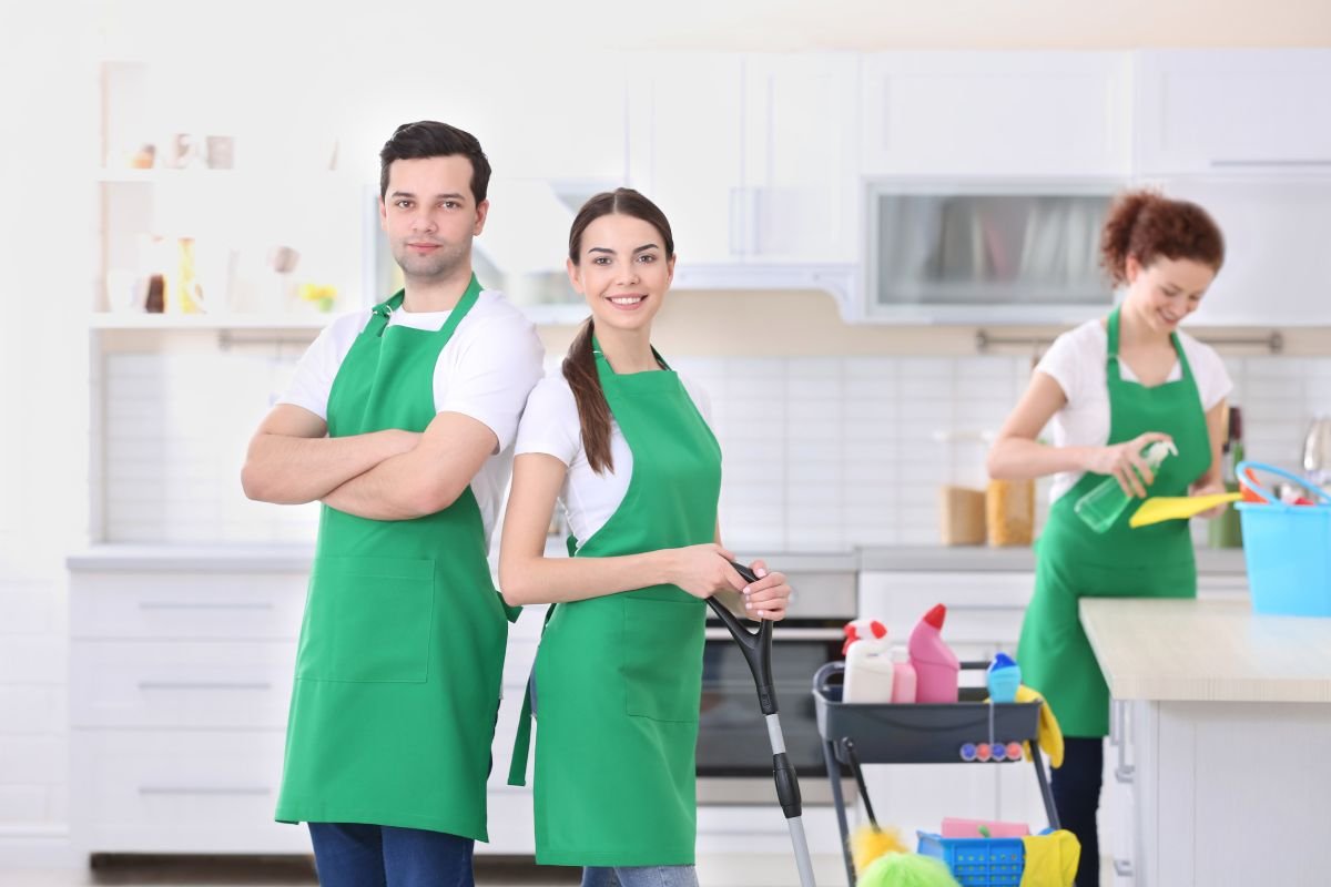 Three individuals wearing green aprons stand together in a kitchen, surrounded by cooking utensils and ingredients.